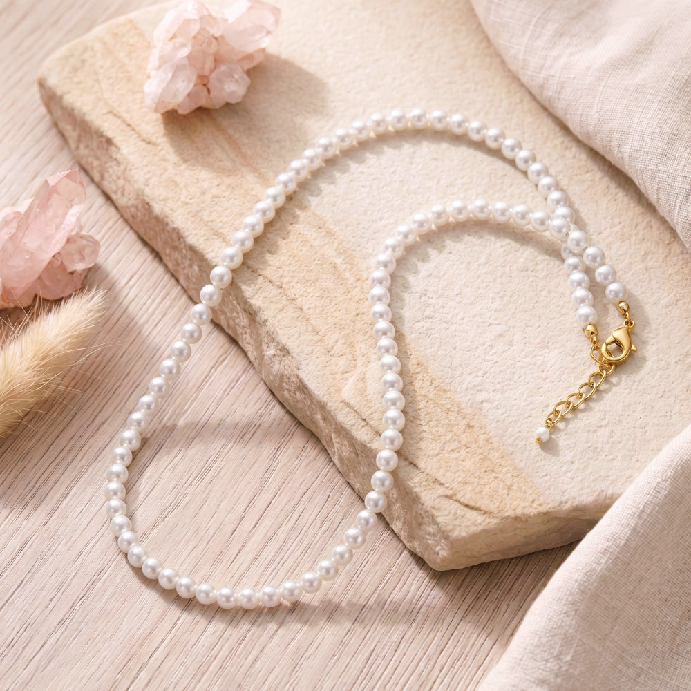 A white pearl necklace with a gold clasp and chain extension, styled on a beige stone slab with soft pink crystals and linen fabric.