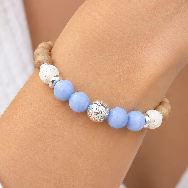 A close-up of a beaded bracelet worn on a wrist. The bracelet features light blue round beads, two white textured beads, a central metallic silver bead with a floral pattern, and natural wood beads at the ends.