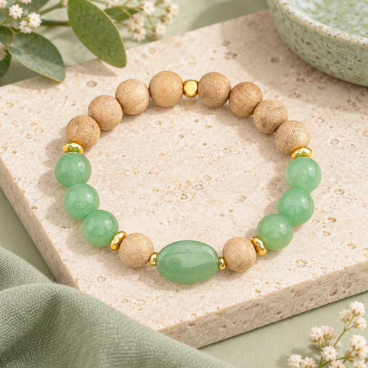 A beaded stretch bracelet featuring light green jade-like round beads, natural wood round beads, and small gold spacer beads, displayed on a textured beige stone slab with soft green fabric and floral props.